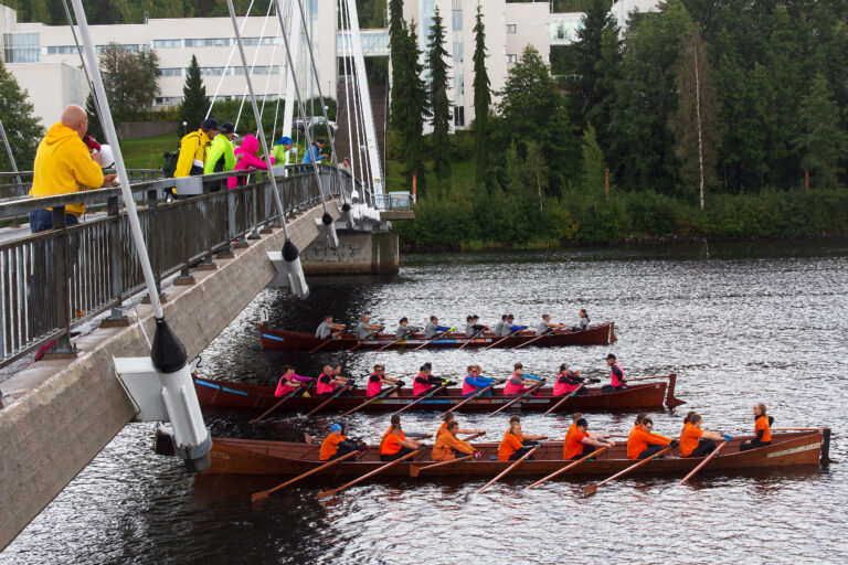 Kolme kirkkovenettä täynnän ihmisiä lähdössä soutamaan Jyväsjärven syksyisessä ilmassa.